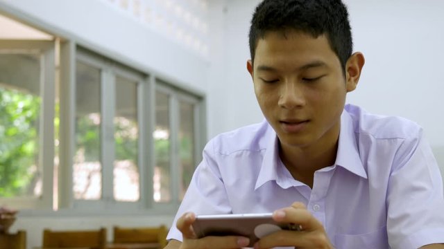 Asian male high school student in a white uniform, who is a lot of addicted to games. He is playing exciting games on  their mobile phones and sitting in the classroom.