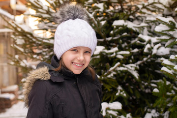 The young girl smiles charmingly in winter, she stands in a warm hat and jacket near a large Christmas tree, there is a lot of snow everywhere