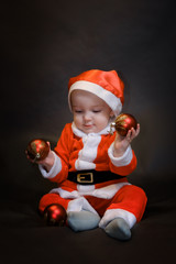Little baby dressed as Santa Claus sits on a dark background and plays with Christmas toys