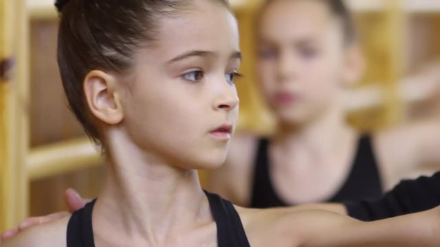 Close up faces of concentrated young girls of primary school age standing by wall bars and practicing ballet movements, handheld