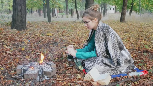 The yong caucasian woman in a warm green sweater sitting by the fire in the evening in foreat at autumn.