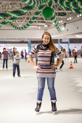 Obraz premium Portrait of cheerful woman dressed sweater, jeans and scarf standing on ice in hockey skates, indoor ice-scating rink with people