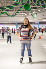Portrait of cheerful woman dressed sweater, jeans and scarf standing on ice in hockey skates, indoor ice-scating rink with people