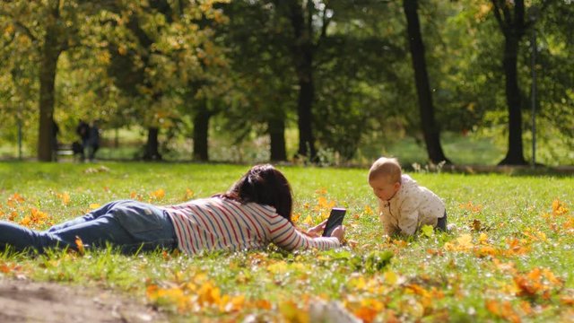 Mother lie on a park grass takes phone picture of baby child toddler daughter try crawling towards her