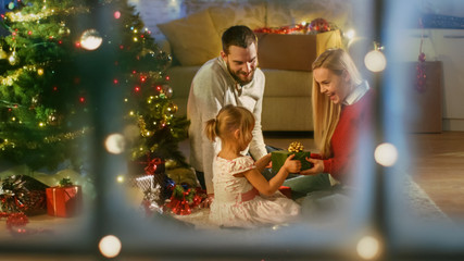 Looking Through Snowy Window. Happy Father, Mother and Daughter Sitting Under Christmas Tree. Daughter Gives a Gift to Her Mother.