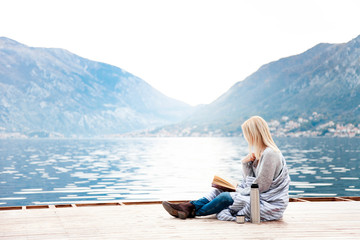 Woman is reading on wooden pier by winter sea, beach, mountains. Cozy picnic with coffee, hot beverages, tea in thermos and mug, warm plaid, opened book. Girl is enjoying nature, wellbeing.