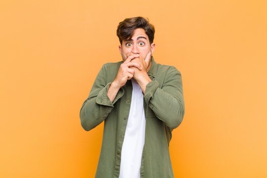 young handsome man covering mouth with hands with a shocked, surprised expression, keeping a secret or saying oops against orange background
