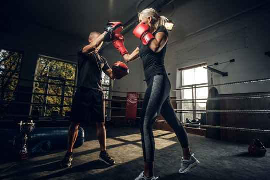 In the dark gym on the ring experienced trainer and young woman has a kick boxing fight.