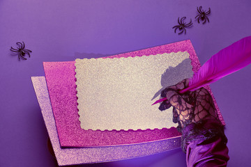 Creative purple and pink Halloween background with copy-space. Hand in black mesh glove holding pink quill over stack of glittering paper on purple background with spiders