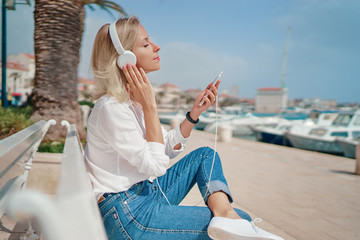 Enjoying the sound. Happy young woman with earphones listening music on smartphone while sitting on city embankment sea promenade.