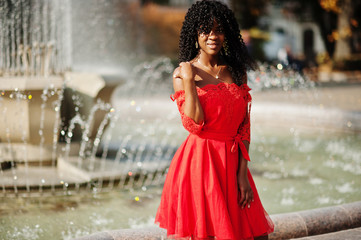 Stylish trendy afro france curly woman posed at autumn day in red dress. Black african female model against fountain.