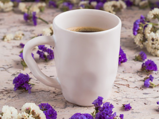 Сeramic white cup of coffee with dry white and violet flowers on the light background. Morning americano. Cafe and bar, barista art concept.