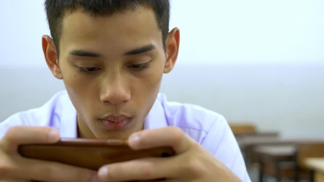 An Asian male high school student in a white uniform, who is a lot of addicted to games, is playing exciting games on his mobile phone and sitting on the school stairs.