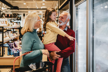 Happy grandmother and grandfather with granddaughter shopping together in grocery store or...