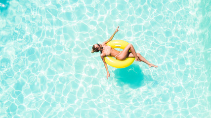 Top view of slim young woman in bikini on the yellow air inflatable ring in the big swimming pool.
