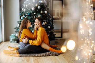 Loving couple, is stylishly dressed, sitting on a rug and hugging each other near a Christmas tree. Christmas time. Christmas mood