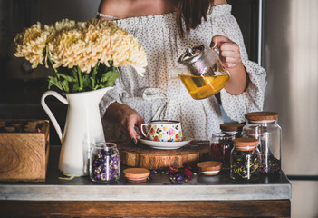 Young caucasian woman pouring freshly brewed green tea from glass pot into beautiful vintage porcelain cup at kithcen counter. Autumn tea time concept