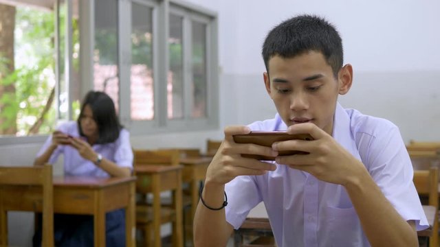 Asian high school students in a white uniform, who are a lot of addicted to games. They are playing exciting games on  their mobile phones and sitting in the classroom.