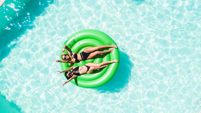 Aerial view of two women lying on inflatable green mattress floating and relaxing in pool