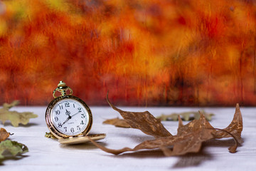 Golden pocket watch and fall leaves on white wooden table at wet window glass. Autumn time concept.