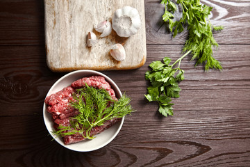 Bowl with raw minced meat, cutting board with fresh seasonings on a wooden background. White garlic heads and cloves, green dill and parsley leaves. Ground beef and condiments, top view