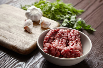 Bowl with raw minced meat, cutting board with fresh seasonings on a wooden background. White garlic heads and cloves, green dill and parsley leaves. Ground beef and condiments