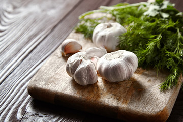 Cutting board with fresh seasonings on a wooden background. White garlic heads and cloves, green dill and parsley leaves. Condiments