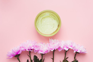 Green aloe gel in a jar on a pink pastel background next to a chrysanthemum flowers. The concept of skin hydration, skin care, natural ingredients and anti-aging. Sunburn protection, wound healing