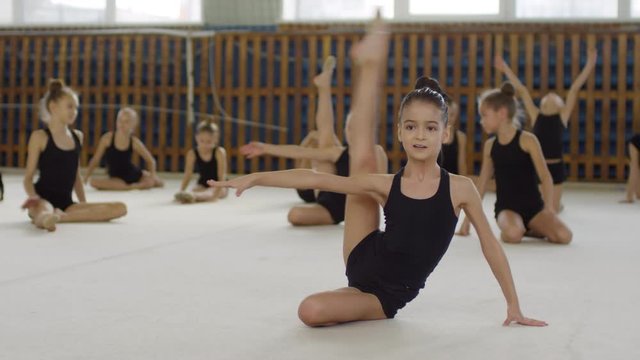 Young girl of elementary school age wearing sports clothes and ballet slippers, performing leg stretching exercises and looking at camera