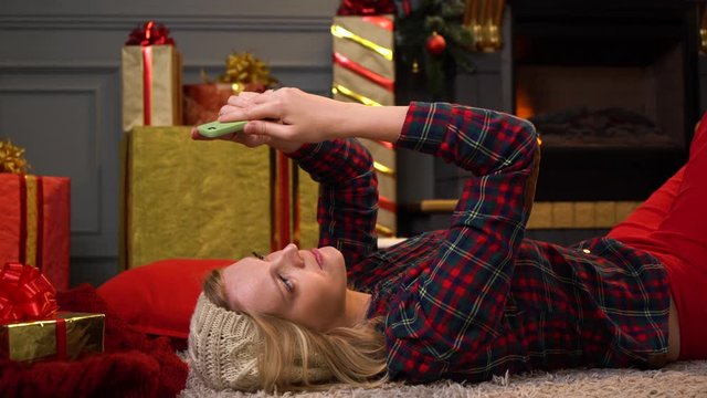 Young beautiful girl or woman lie on floor in a red checkered shirt taking video call by the phone with christmas tree on background.