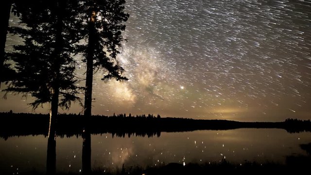 A dusk to dawn star trails time lapse of the Milky Way with pine tree silhouettes in the foreground.  A large lake shows reflections of the stars.  Weak Aurora occur at the end of the video.