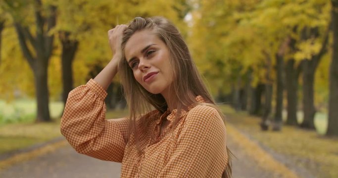Profile portrait of a confident Caucasian woman standing on the road in the autumn park. Charming girl in mustard dress tucking hair behind ear and smiling. Cinema 4k footage ProRes HQ.