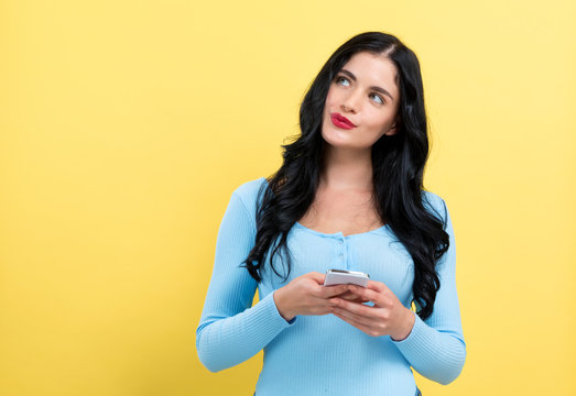 Happy young woman with smart phone thinking about something on a yellow background