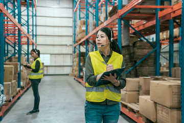Warehouse worker woman wearing headset holding clipboard in large warehouse. blurred view colleague...