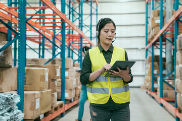 Businesswoman using digital tablet in distribution warehouse. young woman staff in headset talking to customer online while walking in large storehouse. female colleague in background check inventory