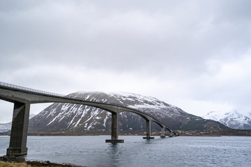 A long concrete bridge for cars and trucks crossing sea under cloudy sky. Snowy mountain on the background and beautiful Lofoten landscape. Atlantic Ocean Road Norway curved concrete bridge and water.