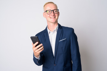 Happy young blonde businessman in suit thinking while using phone