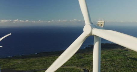 Aerial orbiting close up of a windmill spinning at sunrise with other windmills, green fields, and ocean in the background, wind farm generating electricity, green energy future
