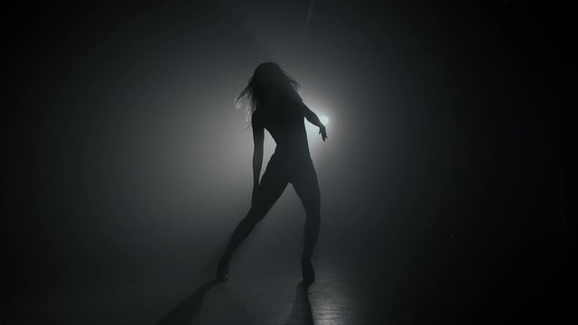 Silhouette of beautiful dancer on smoky dark background.Spotlight shines back against sexy girl. She moving seductively.