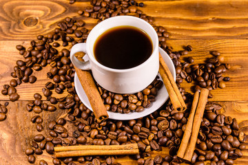 Cup of hot coffee, cinnamon sticks and scattered coffee beans on wooden table