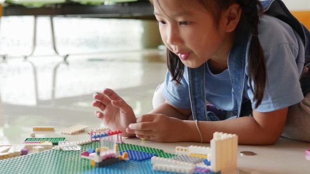 Asian elementary school girl student wearing in light blue T-shirt is lying on the floor and playing creative toys by connecting each parts to another parts together as imaginatively.