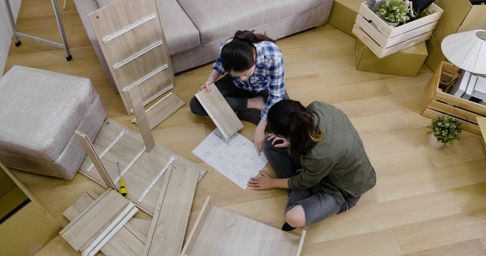 Top View Two Young Lesbian Couple Installing Assembly Furniture In New House With Instructions Paper. Women Reading Diy Manual Together In Living Room. Female Same Sex Marriage Purchase Real Estate