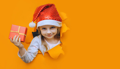 A beautiful little smiling girl in a red Santa hat looks out of a hole in orange paper and holds a box with a gift in her hand. Christmas and New Year concept. Bright coloured background, copy space