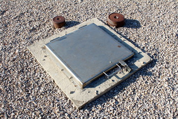 Closed grey heavy metal manhole cover locked with padlock surrounded with concrete foundation and gravel next to two small rusted pipes on warm sunny summer day