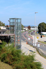 Glass elevator on side of train station surrounded with dense vegetation and street under reconstruction with multiple warning road signs on warm sunny summer day