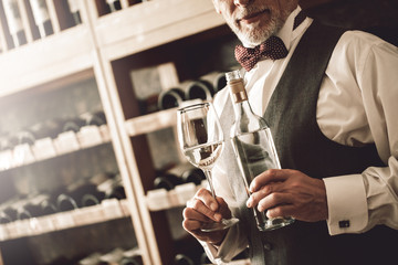 Sommelier Concept. Senior man standing with bottle and glass of white wine smiling confident close-up