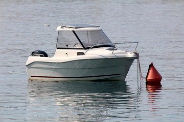 Small fishing boat with modern solar panel on top and inox rail in front tied to large red buoy in local harbor surrounded with calm clear blue sea on warm sunny summer day