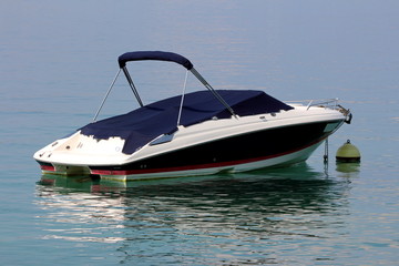 Fototapeta premium Small speed boat covered with dark blue protection anchored in local harbor surrounded with calm clear blue sea on warm sunny summer day