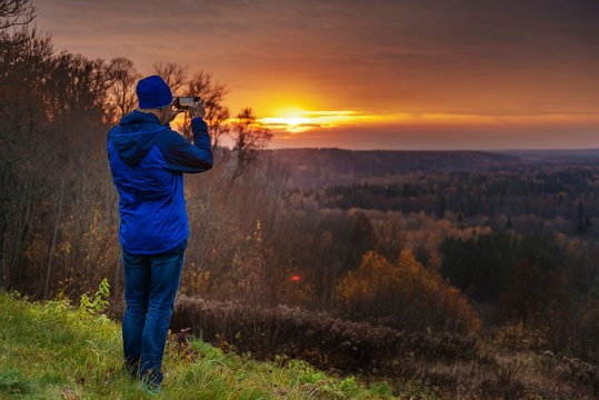 Man in blue taking pictures with a phone of a beautiful sunset.