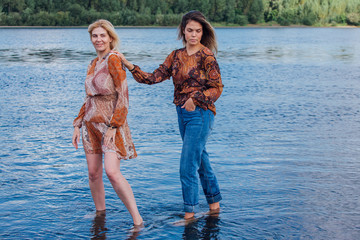 Two pretty sisters standing in river next to the shore on a cloudy windy summer day, having fun and laughing.